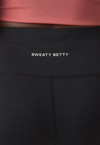 Leggings neri con un alto elastico in vita, con la scritta "SWEATY BETTY" in bianco. Il tessuto sembra liscio e elasticizzato.
