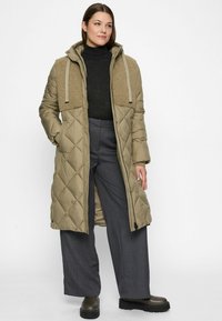 Woman wearing long olive quilted coat with textured shoulder panels, black turtleneck, wide gray pants, and dark boots, standing against white background.