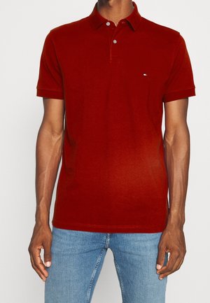 Man draagt een rood poloshirt met korte mouwen en knopen, gecombineerd met lichtblauwe spijkerbroek, staand tegen een effen achtergrond.