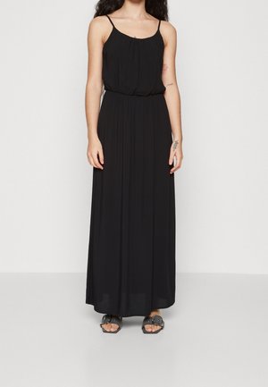 Kobieta w czarnej, bezrękawnikowej sukience maxi z cienkimi ramiączkami i czarnych sandałach na odkryte palce, stojąca na tle jednolitego, jasnego tła.