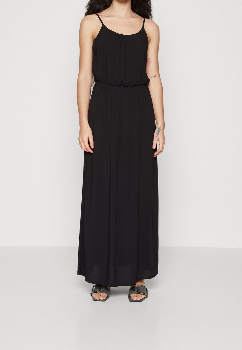Femme portant une robe maxi noire sans manches avec de fines bretelles et des sandales noires à bouts ouverts, debout devant un fond clair uni.