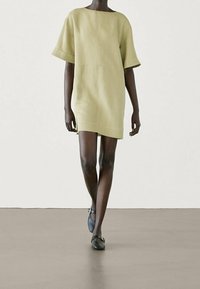 Robe courte en lin vert doux avec une coupe carrée, épaules tombantes et ourlet droit. Portée avec des ballerines noires, sur un fond uni.