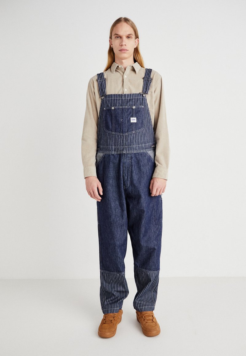 Pantalones de mezclilla en azul oscuro con detalles a rayas, que cuentan con un bolsillo en el pecho y bolsillos laterales, combinados con una camisa beige de botones y zapatos marrón claro.