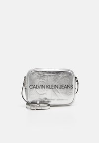 Stříbrná obdélníková krosbody taška s texturovaným povrchem. Obsahuje embosované logo "CALVIN KLEIN JEANS" a nastavitelný popruh.