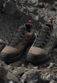 Tan and black hiking shoes feature a mesh upper, textured pattern, and rubber soles. Red accents on the heel and sturdy laces for support.