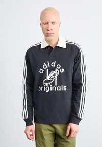 Polo cu mânecă lungă negru cu guler alb și trei dungi albe pe mâneci, având un print grafic cu o încălțăminte și textul "adidas originals."
