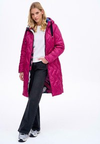 Dlouhý, růžový prošívaný kabát s kapucí, s vzorovanou podšívkou, v kombinaci s bílým topem a černými rozšířenými džíny. Stříbrné sportovní boty.