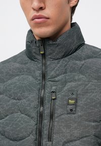 Jachetă puffer gri, matlasată, cu guler înalt, fermoar bidirecțional și patch cu brand. Prezintă un finisaj texturat și un model geometric subtil.