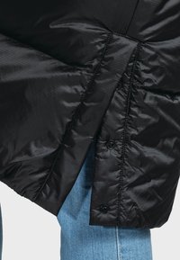 Schwarze Daunenjacke mit glänzender Oberfläche, ausgestattet mit einer Knopfleiste und strukturiertem Muster, kombiniert mit hellblauen Jeans.