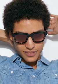 Ochelari de soare cu ramă neagră, având accente din material tip țestoasă, lentile dreptunghiulare și nuanță gradient. Purtați împreună cu o cămașă din denim.