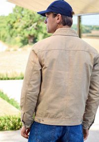 Man wearing tan jacket and blue cap stands outdoors facing away, with greenery and a field in the background.