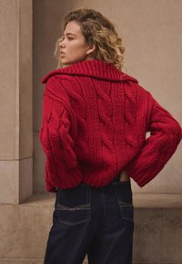 Red cable-knit sweater with a large collar, featuring chunky texture and oversized sleeves, paired with dark denim jeans.