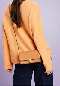 Jasnobrązowa torba na ramię z złotym zapięciem, prostokątna forma, gładka tekstura, noszona w zestawieniu z pomarańczowym swetrem i ciemnymi dżinsami.