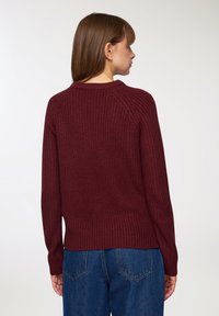 Bordowy sweter dzianinowy z teksturowanym wzorem, ściągaczami przy mankietach i u dołu oraz rękawami raglanowymi. Noszony z niebieskimi dżinsami o wysokim stanie.