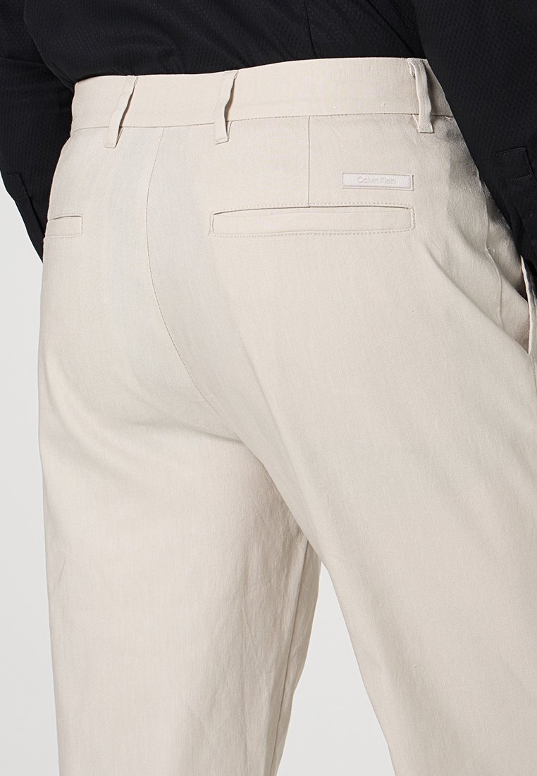 Beige maßgeschneiderte Hose mit Gesäßpaspeltaschen und Gürtelschlaufen, getragen mit einem schwarzen Langarmoberteil.