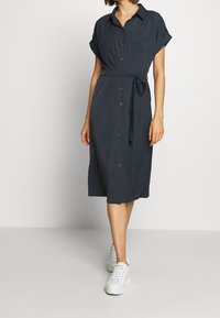 Abito camicia blu navy con maniche corte arrotolate, cintura in vita e tasche frontali, abbinato a sneakers bianche. Tessuto morbido e lunghezza media.