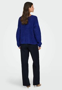 Prevelik modri pulover iz teksturirane tkanine, v kombinaciji z temnimi širokimi hlačami in bež petkami. Pulover ima sproščen kroj in dolge rokave.