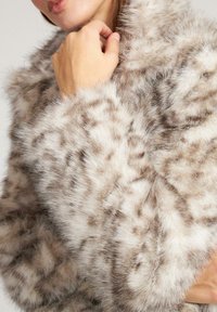 Ekskluzywny szal z futra sztucznego w jasnobeżowym kolorze z ciemniejszymi plamkami. Miękka tekstura z szerokim kołnierzem i obszernymi mankietami, zapewniająca przytulny wygląd.