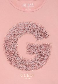 Svijetlo ružičasta majica od organskog pamuka s teksturiranom "G" koja ima latice i zlatne detalje, s natpisom "GUESS EST. 1981" otisnutim ispod.