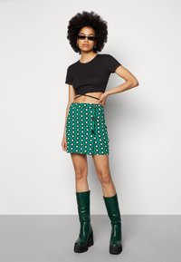 Top cropped preto canelado combinado com uma saia mini verde com padrão que apresenta flores brancas e azuis, e botas pretas de cano alto com plataforma.