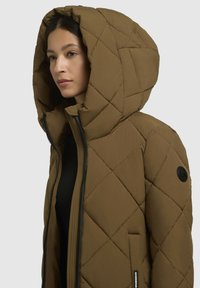 Giacca a vento trapuntata marrone con cappuccio oversize, dotata di una chiusura frontale con zip, tasche laterali e un'etichetta rotonda con logo sulla manica.