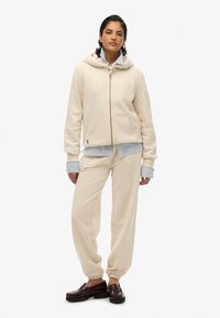 Sweatshirt à capuche beige avec une fermeture éclair à l'avant et des poches, assorti à un pantalon de survêtement. Chemise rayée sous le hoodie et mocassins foncés.