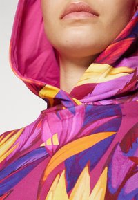Närbild av en person som bär en färgstark huvtröja med gula, rosa, lila och orangea bladmönster, som visar nedre delen av ansiktet och nacken.