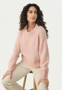 Pull en tricot rose avec un col rond, des poignets côtelés et un ourlet. Porté avec un pantalon en velours côtelé beige. Modèle assis sur un tabouret en bois.