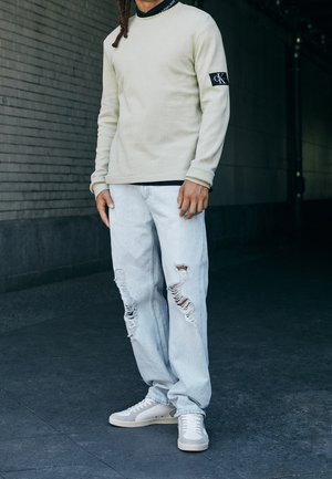 Osoba ubrana w kremową bluzkę z długim rękawem z logo Calvin Klein na rękawie, jasnoniebieskie porwane dżinsy oraz białe trampki, stojąca na ciemnym chodniku.