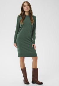 Zaļa adīta kleita ar garām piedurknēm, ribota apkakle un malu. Apvienota ar brūnām ādas augstiem zābakiem; gluda tekstūra un pieguļoša forma.