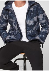 Marinblå jacka med ett grått abstrakt mönster, dragkedja fram, snoddar på huvan och två sidofickor. Bärs över en vit T-shirt.