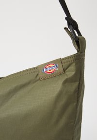 Olivno zelena torba iz teksturirane tkanine s pravokotno obliko. Ima izšiteno oznako logotipa in črno nastavljivo naramnico.