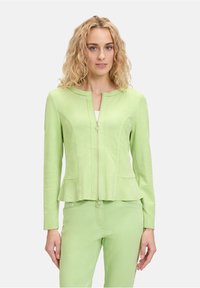 Jasnozielona kurtka zapinana na zamek, z fasonem peplum, gładką teksturą i dwoma przednimi kieszeniami. Stylizowana z dopasowanymi zielonymi spodniami.