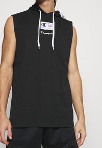 Black sleeveless hoodie made of cotton, featuring white drawstrings, a hood, and a logo patch on the chest. Smooth texture, casual design.