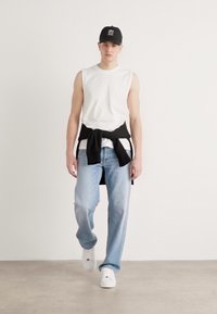 Camisa sin mangas blanca, sudadera negra atada a la cintura, jeans azul claros y zapatillas blancas. Una gorra negra completa el look casual.