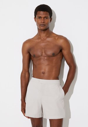 Muskelbepackter Mann steht an einer weißen Wand, oberkörperfrei, trägt hellbeige Shorts mit der Hand in der Tasche, schaut mit neutralem Gesichtsausdruck nach vorne.