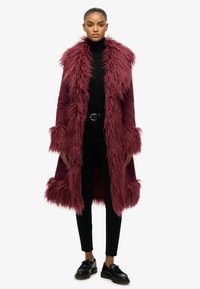 Bordeaux bontjas met lange mouwen, pluche kraag en manchetten, met een zachte textuur en een middellange ontwerp. Draagt boven een zwarte outfit.