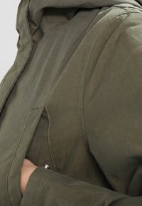 Olivově zelená texturovaná látková bunda s kapucí, viditelným šitím a přední kapsou, se stříbrným patentním knoflíkem.