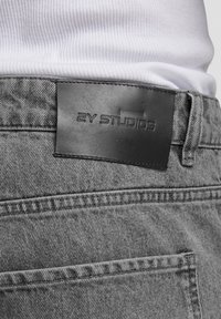 Graue Jeans aus Denim mit strukturiertem Finish, ausgestattet mit einem schwarzen Lederetikett, das am Bund mit "2Y STUDIOS" geprägt ist.