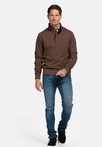 Man loopt en draagt een bruine trui met kwartrits over een gestreept shirt, blauwe jeans en zwarte sneakers op een witte achtergrond.