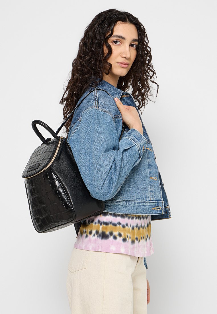 Zaino in pelle nera con stampa a coccodrillo e finitura testurizzata. Presenta spallacci regolabili e chiusura con zip. Indossato con una giacca di denim.