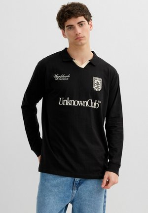 Joven con polo de manga larga negra con el texto "UnknownClub" y escudo, combinado con jeans azul claro, posando contra un fondo liso.