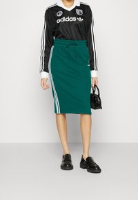 Musta pitkähihainen pelipaita valkoisilla yksityiskohdilla yhdistettynä petroolin väriseen pelipaitahameeseen, jossa on valkoiset sivuraidat, viimeistelty mustilla loafereilla ja käsilaukulla.