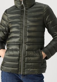 Giacca a vento verde con cuciture orizzontali, colletto alto, zipper frontale e tasche laterali, realizzata in materiale imbottito e lucido.