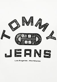 Λευκό ύφασμα με έντυπο σε έντονο, ταλαιπωρημένο μαύρο χρώμα, που γράφει "TOMMY JEANS" και έχει ένα κεντρικό γραφικό με αλτήρα, με την ημερομηνία "1985" από κάτω.