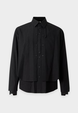 Homme portant une chemise noire à boutons superposée, un pantalon gris ample et des chaussures en cuir rouges avec des lacets rouges, debout devant un fond blanc uni.