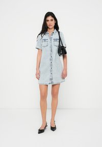 Φόρεμα από ανοιχτό μπλε τζιν με κοντά μανίκια, κουμπιά μπροστά και τσέπες στο στήθος. Διαθέτει ξεφτισμένο ζωνάρι και συνδυάζεται με μαύρη τσάντα και παπούτσια.