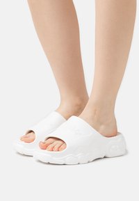 Sandalias de diapositiva blancas con un upper texturizado, tira ancha y diseño de punta abierta. Suela gruesa con un perfil ondulado y detalle sutil del logo.