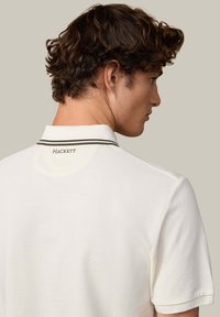 Biele polotričko s textúrovanou látkou, s kontrastným prúžkovaným golierom a vyšívaným logom "HACKETT" na zadnej strane krku.
