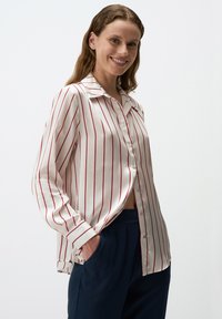 Camicia bianca con sottili righe rosse, colletto e maniche lunghe; abbinata a pantaloni blu navy, in tessuto liscio e dalla vestibilità ampia.